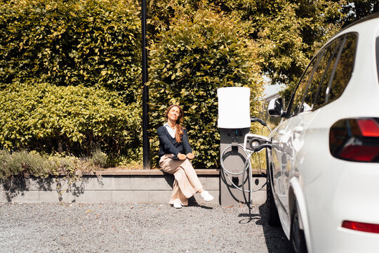 Businesswoman relaxing by electric car at fast charging station outdoors
