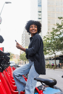Smiling businesswoman holding smart phone sitting on electric bicycles arranged in rows