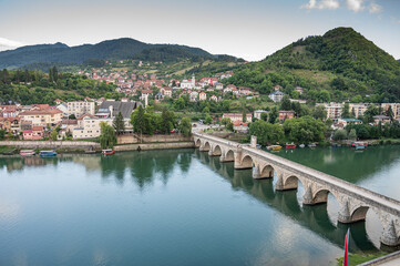 An old stone bridge with arches spans the tranquil Drina River, surrounded by green hills