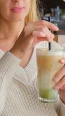 Woman sipping iced matcha latte with straw while sitting in cozy cafe wearing white sweater