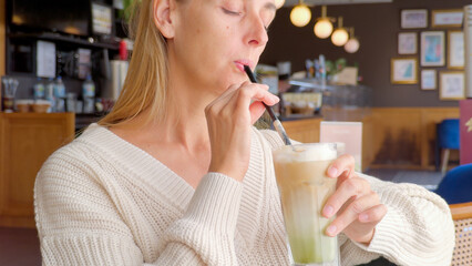 Woman sipping iced matcha latte with straw while sitting in cozy cafe wearing white sweater