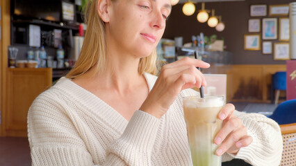 Woman stirring iced matcha latte with straw while sitting in cafe wearing white knitted sweater