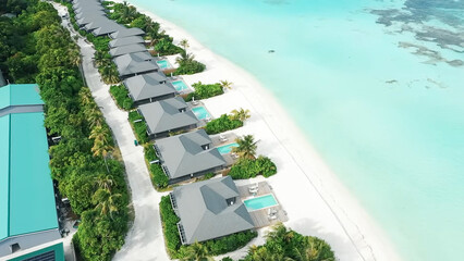 An aerial view of a row of luxury beachfront villas, each with a private pool, lining the pristine white sand of the ocean shore