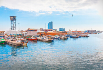 View of Port Vell in Barcelona, featuring docked small boats, historical waterfront buildings, and the distinctive Torre Jaume I in the background. The city skyline is visible under a cloudy sky