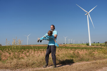 A happy father carries his smiling son in the air with a windmill and a sky with clouds in the background.