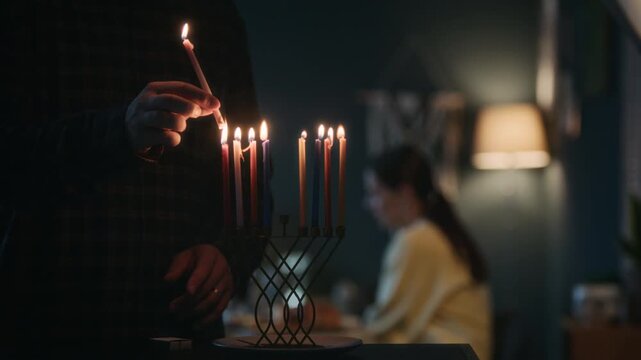 Young Jewish man lighting up candles on hanukkiah with shammash and then joining his wife and toddler son at heartwarming festive table, celebrating Hanukkah with family at home