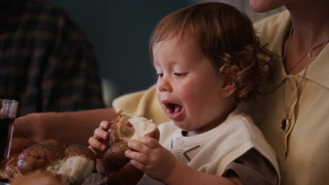 Close up shot of adorable toddler boy sitting on parents lap and enjoying traditional bread challah celebrating Jewish holiday with family at home
