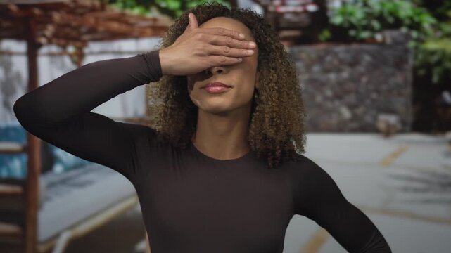 Transgender woman with curly hair covering eyes with hand on urban street by stone wall and wooden bench; vulnerability.