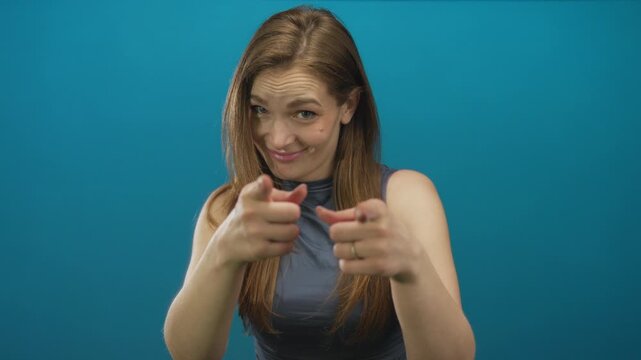 Young caucasian woman finger pointing with both hands in blue studio setting while smiling confidently; playfulness motivation encouragement positivity.