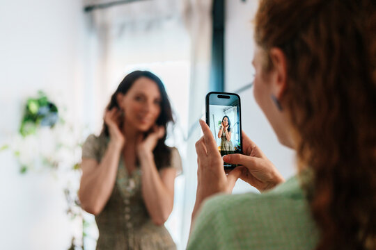 Woman taking photo of another woman posing with smartphone at home