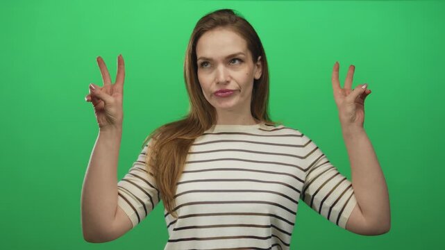 Young caucasian woman wearing striped top raises both hands to form air quotes gesture in green studio; sarcasm.