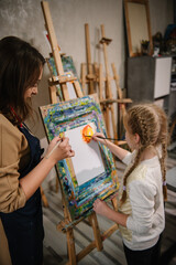 School girl drawing brush landscapes on easel. Teacher explains to kid how to draw pictures in art class. Child enjoys painting in workshop. Art lesson. Education, creativity, learning concept.