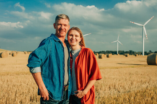 Cowboy and partner smiling in wheat field with wind farm and hay bales - Powered by Adobe