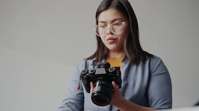 Low angle shot of concentrated brunette woman holding professional camera and adjusting props on table while shooting in modern minimalist studio