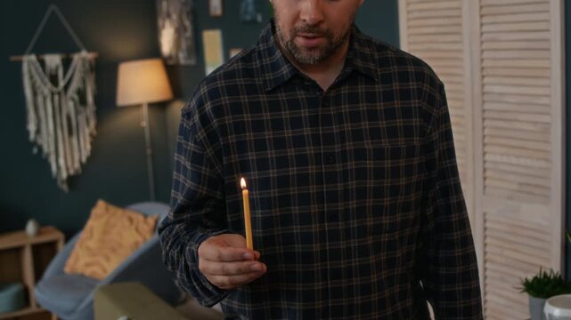 Tilt down shot of young Jewish man holding burning shammash and lighting up candles on hanukkiah while singing holiday hymn Maoz Tzur, celebrating Hanukkah at home