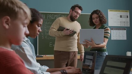 Medium shot of teenage Caucasian schoolgirl demonstrating coding project to young male teacher, who is checking and giving feedback, while students are writing programs on laptops during lesson - Powered by Adobe