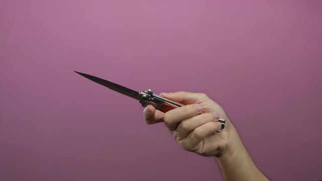 Caucasian man's hand holding a knife against a pink background, highlighting the sharp blade and wooden handle, creating an isolated and focused visual on the weapon.