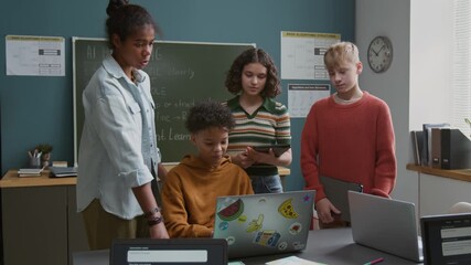 Medium shot of four diverse teenage schoolkids working on project together in classroom during programming lesson, biracial boy writing computer code on laptop while African American girl commenting - Powered by Adobe