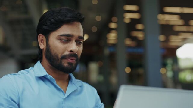 Portrait of happy Indian entrepreneur gestures on video call via laptop in street cafe. Cheerful man uses sign language during online meeting using device outside corporate office