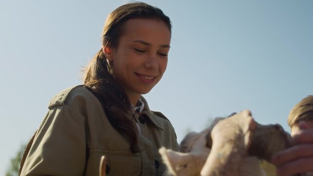 Young cheerful woman smiling gently stroking goat while tending livestock on farm or attending petting zoo captured against early autumn sky