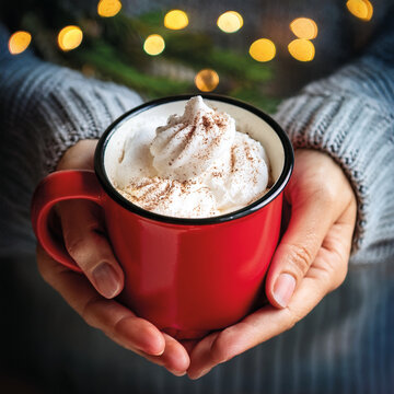 Hands holding a red mug with hot cocoa and whipped cream, wearing wool sweater, blurred Christmas lights background, cozy winter mood