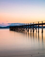 A long wooden pier stretches out over calm, reflective water toward the horizon at sunset. Gentle hues of orange and purple grace the sky