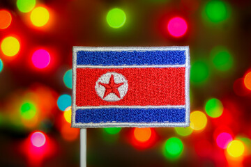 Handmade embroidery of the North Korea flag (DPRK) with red star and circle. Set against a vibrant, creamy celebration bokeh background. Ideal for national holidays or Asian political themes.