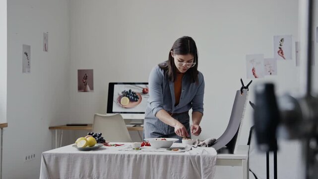 Brunette food stylist adjusting plates, cleaning rim of plate with brush while preparing props for shooting