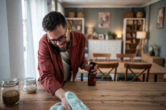 Happy man cleaning wooden table while talking on phone
