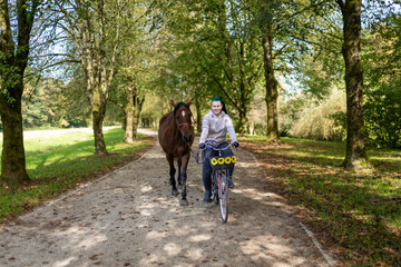 Obraz premium Young Woman Riding Bicycle Along Tree Lined Path Leading Brown Horse on Sunny Day in Nature