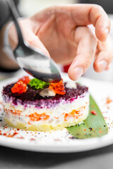 Layered gourmet salad with beetroot, potato, carrot, and mayonnaise, garnished with red caviar and parsley, served on a white plate. Close-up of hand adding final touch.