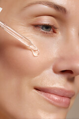 Close-up of woman applying acid serum with glass dropper on cheek, glowing skin, soft diffused light, neutral spa background.