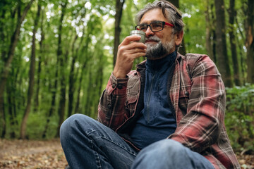 Fototapeta premium Enjoying the hot drink from thermos. Hiking experience, senior man is in the forest at daytime