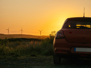 Fototapeta premium A red car is parked in a field with a sunset in the background. The car is facing the sun, and the sky is orange and pink. Wind farm turbines in the background. Sightseeing modern technology concept
