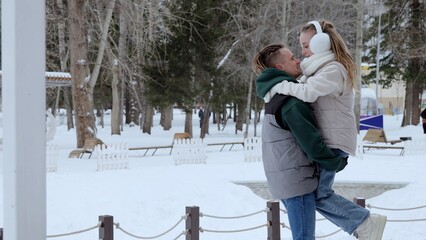 Romantic couple embracing, male partner lifting female partner amid snow covered park landscape, sharing intimate winter moment together