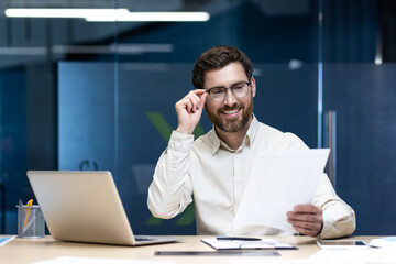 Smiling young male businessman working in office with documents, reading papers and holding glasses in hand