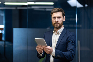 Portrait of a young serious businessman in a suit standing in the office, holding a tablet and looking at the camera