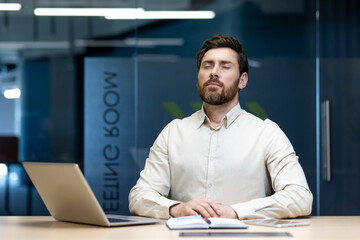 A young man sits in the office at his workplace, his eyes closed and resting