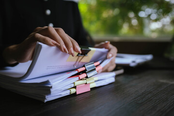 Businessperson organizing and reviewing stacks of paper documents and reports on a wooden desk in an office environment.