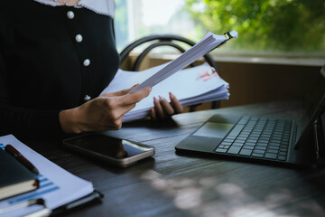 Businessperson organizing and reviewing stacks of paper documents and reports on a wooden desk in an office environment.