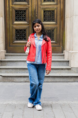 A teenager in a red jacket and blue jeans stands on a sidewalk