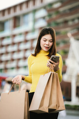 A cheerful young woman in a yellow sweater holds paper shopping bags and a smartphone, enjoying a fun shopping day outdoors.