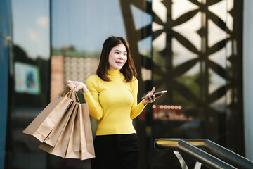A cheerful young woman in a yellow sweater holds paper shopping bags and a smartphone, enjoying a fun shopping day outdoors.