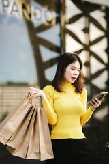 A cheerful young woman in a yellow sweater holds paper shopping bags and a smartphone, enjoying a fun shopping day outdoors.