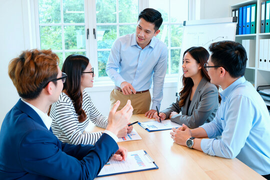 Group of young adult Asian men and women discussing business strategy around meeting table, one man standing and presenting while others listening and taking notes during team meeting