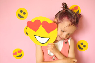 A beautiful shy sweet girl with dyed pink hair holds a smiley face reflecting love. Emojis with hearts instead of eyes. World Social Media Day