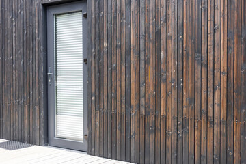 A modern wooden wall with a sleek glass door. The wood has a dark finish, showcasing a contemporary architectural style. The door features horizontal blinds.