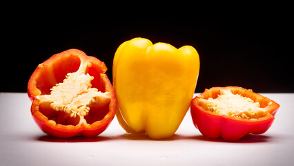 A vibrant still-life studio photograph showcasing a whole yellow bell pepper placed between two halved red bell peppers with visible seeds, set on a clean white surface against a contrasting black bg