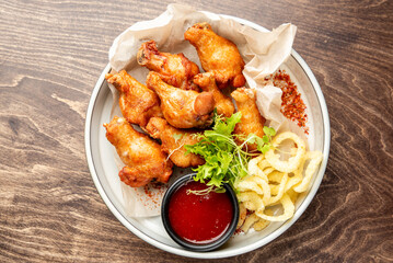 Crispy fried chicken drumsticks with curly fries, red dipping sauce, and fresh herbs on parchment paper over a white plate. Rustic wooden table background, appetizing fast food presentation.