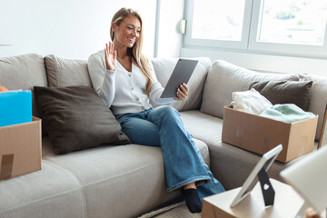 Obraz premium Woman using tablet PC sitting on sofa in living room at new home. Young woman surrounded with unpacked cardboard boxes sitting on the sofa in her new apartment and using digital tablet.